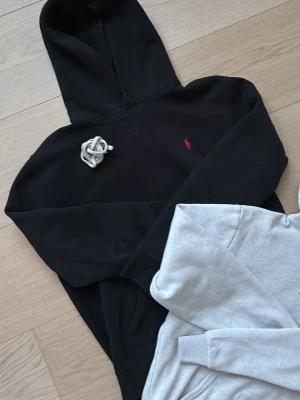 Svart hoodie från Polo Ralph Lauren - Säljer en svart hoodie från Polo Ralph Lauren med den klassiska röda loggan broderad på bröstet. Hoodien har huva och magficka, samt ribbade muddar vid ärmslut och nederkant. Perfekt för en avslappnad och stilren look.