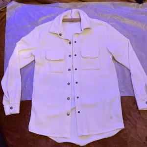 Vit overshirt med tryckknappar - Stilren vit overshirt med långa ärmar och två bröstfickor med lock. Skjortan har tryckknappar framtill och på ärmsluten samt en klassisk krage. Perfekt att bära öppen eller stängd för en avslappnad look.