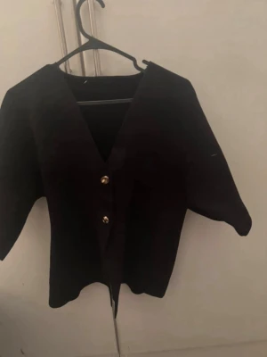 Svart kofta med guldknappar - Stilren svart kofta med djup V-ringning och två dekorativa guldknappar framtill. Koftan har korta ärmar och rak passform, perfekt för att lyfta en enkel outfit. Materialet ser ut att vara mjukt och bekvämt. För mer bilder/justering av pris skriv gärna❤️🤗