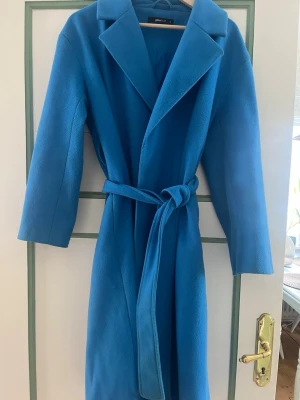 Blå kappa med knytskärp från Gina Tricot - Stilren blå kappa från Gina Tricot med bred krage och knytskärp i midjan. Kappan har en rak passform och är tillverkad i ett mjukt tyg som ger en snygg siluett. Perfekt för dig som vill ha en färgklick i garderoben.