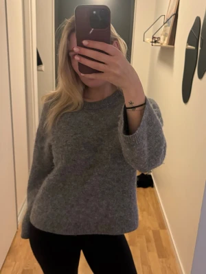 Grå stickad tröja från Arket - Mysig grå stickad tröja med rund hals och långa ärmar. Tröjan har en avslappnad passform och är tillverkad i ett mjukt, fluffigt material som passar perfekt för kyligare dagar. Enkel och stilren design med bra kvalité.