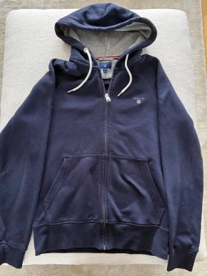 Mörkblå zip hoodie från GANT - Snygg mörkblå hoodie från GANT med dragkedja, vit dragsko och grått foder i huvan. Klassisk GANT-logga broderad på bröstet och stora kängurufickor framtill. Perfekt för en avslappnad och stilren look.