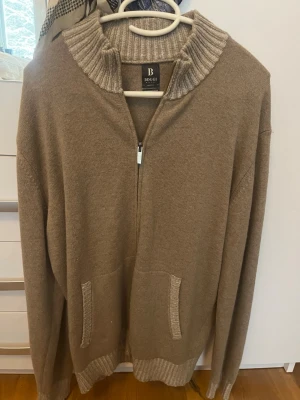Beige stickad tröja från Boggi - Stickad tröja i beige från Boggi med full zip. ribbad hög krage och muddar. Tröjan har två fickor framtill och är tillverkad i mjukt material, perfekt för lager på lager. Enkel och stilren design som passar till det mesta. NEVER WORN.