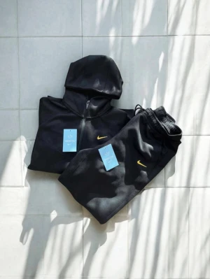 Svart Nike tracksuit med huva - Säljer en svart tracksuit från Nike med gul logga. Setet består av en hoodie med dragkedja och två fickor samt matchande byxor med resår i midjan. Perfekt för träning eller chill. Materialet är mjukt och stretchigt för maximal komfort.