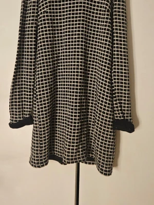 Svartvit rutig vintage klänning - Mönstrad svartvit klänning med långa ärmar. Klänningen är figursydd och är tillverkad i ett mjukt, stickat material. Perfekt för dig som gillar grafiska mönster och en avslappnad stil.