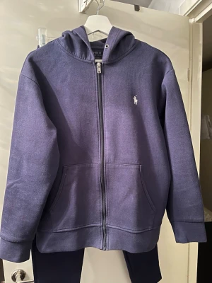 Mörkblått set från Polo Ralph Lauren - Snyggt mörkblått set från Polo Ralph Lauren med hoodie och matchande byxor. Hoodien har dragkedja, fickor och logga på bröstet. Byxorna har resår i midjan, fickor och logga på höften. Båda i mjukt bomullsmaterial för en avslappnad look.