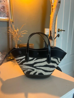 Zebramönstrad handväska med läderdetaljer - Snygg handväska med zebramönstrad textil och guldiga detaljer. Väskan har en unik, bred form och både handtag och axelrem för olika stilar. Perfekt för dig som vill sticka ut med ett djurmönster och trendig design.