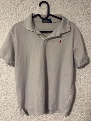 Ljusgrå piké från Polo Ralph Lauren - Klassisk ljusgrå pikétröja från Polo Ralph Lauren med korta ärmar och krage. Tröjan har den ikoniska röda logotypen broderad på bröstet och är tillverkad i mjuk bomull. Perfekt till jeans eller shorts för en clean och stilren look.                               Storlek L men passar bättre M