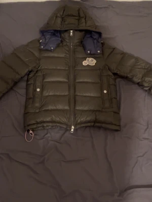 Svart dunjacka från Moncler med huva - Snygg svart dunjacka från Moncler med avtagbar huva och blått innerfoder. Jackan har två framfickor med tryckknappar, dragkedja framtill och Moncler-logga på bröstet. Perfekt för kalla vinterdagar och riktigt stilren look.