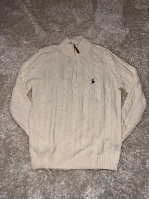Vit kabelstickad tröja Polo Ralph Lauren - Kabelstickad vit tröja från Polo Ralph Lauren med halv dragkedja och ribbad krage. Tröjan har det klassiska mörkblå logobroderiet på bröstet och är tillverkad i mjukt material. Perfekt för en stilren och avslappnad look.