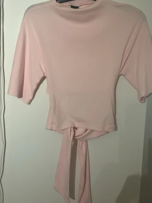Ljusrosa topp med knyt - Supersöt ljusrosa croppad topp med knyt i midjan och korta ärmar. Mjuk och stretchig känsla, troligtvis i bomull eller polyesterblandning. Passar perfekt till både jeans och kjol. Använd 1 gång!