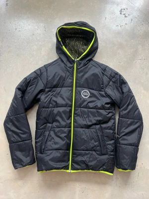 Vändbar svart och mönstrad pufferjacka Wedze - Säljer en pufferjacka från Wedze med limegröna detaljer och huva. Jackan är vändbar med ena sidan helsvart och den andra med mönster i svart och lime. Dragkedja och muddar i lime. Storlek 153-160 cm, 14 år