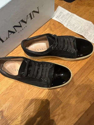 Svarta Lanvin sneakers med Kvitto och Box - Snygga svarta sneakers från Lanvin med klassisk låg silhuett. Skorna har ovandel i mocka och en glansig lackad tå, samt vita sulor som ger en clean kontrast. Perfekta för dig som vill ha en stilren och exklusiv sneaker med unika detaljer. Skorna är köpta på NK och kvitto finns. Orginalbox medföljer ävenz