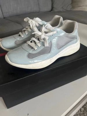 Prada cups  - Snygga ljusblå sneakers från Prada med vita och grå meshdetaljer. Skorna har vita snören, vit sula och en klassisk låg siluett. Materialmix av läder och mesh ger en modern vibe. Perfekta för dig som vill ha något stilrent och trendigt.