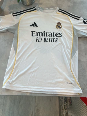 Real Madrid matchtröja Adidas vit - Säljer en vit Real Madrid matchtröja från Adidas med svarta och gula detaljer. Tröjan har klubbmärke och Adidas-logga på bröstet samt 'Emirates Fly Better' tryckt framtill. Tillverkad i lätt och ventilerande material, perfekt för träning eller match.