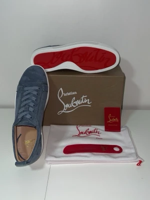 Blåa sneakers från Christian Louboutin - Snygga blåa sneakers i mocka från Christian Louboutin med klassisk röd sula och vita kanter. Skorna har rund tå, matchande blå skosnören och diskret logga på innersulan. Perfekta för dig som vill ha en exklusiv och stilren look.