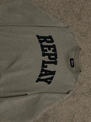 Grå sweatshirt från Replay - Snygg grå sweatshirt från Replay med stor svart logga över bröstet. Tröjan har rund halsringning, ribbade muddar med vita ränder och en patch på ena ärmen. Perfekt för en avslappnad och trendig stil.