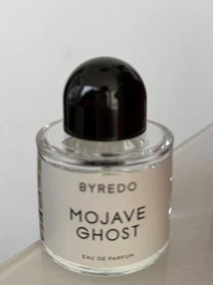 Byredo Mojave Ghost Eau de Parfum - Byredo Mojave Ghost Eau de Parfum i en stilren, rund glasflaska med svart rund kork. Flaskan är transparent med vit etikett och svart text. En modern och exklusiv parfym som passar dig som gillar unika dofter.