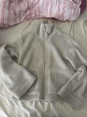 Krämvit teddyjacka med dragkedja - Mysig krämvit teddyjacka med hög krage och dragkedja framtill. Jackan har två stora fickor framtill och är gjord i fluffigt teddy-material som ger en avslappnad och trendig look. Perfekt för kyliga dagar och enkel att matcha med olika outfits.