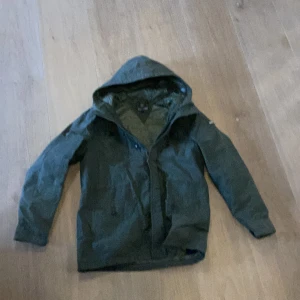 Grön vindjacka från Tommy Hilfiger - Snygg grön vindjacka från Tommy Hilfiger med huva och dragkedja framtill. Jackan har en minimalistisk design och är tillverkad i ett lätt, vindtätt material som passar perfekt för blåsiga dagar. Klassisk passform och diskret logga på ärmen.