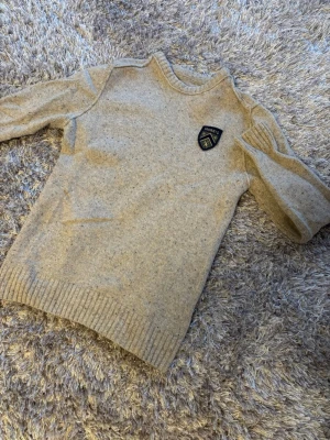 Beige stickad tröja från Morris - Stickad tröja i beige melerad ull från Morris med rund halsringning och ribbade muddar. Tröjan har en snygg patch med Morris-logga på bröstet och är tillverkad i 100% merinoull. Perfekt för dig som gillar klassisk och stilren design. Storlek m Men sitter som S 