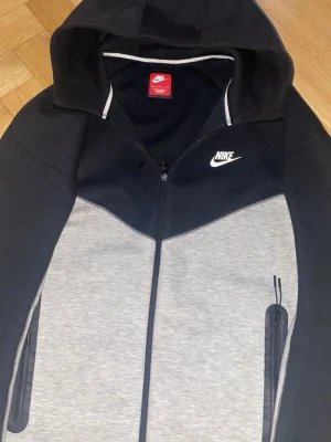 Nike Tech Fleece hoodie svart/grå - Nike Tech Fleece hoodie i svart och grått med dragkedja framtill och två sidofickor med dragkedja. Ikonisk Nike-logga på bröstet och huva med kontrastfärgad kant. Tillverkad i mjukt och bekvämt material som passar perfekt till streetwear.