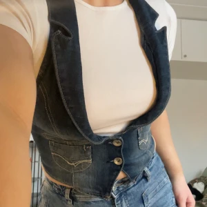 Mörkblå denimväst med fickor - Mörkblå denimväst med två framfickor och dekorativa sömmar. Västen har två knappar framtill och en figurnära passform. I ryggen finns en snörning i korsettstil. Perfekt att bära över en t-shirt för en cool och avslappnad stil. Köpt second hand