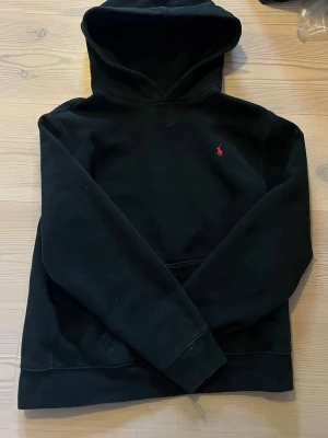Svart hoodie från Polo Ralph Lauren - Klassisk svart hoodie från Polo Ralph Lauren med röd broderad logga på bröstet. Hoodien har känguruficka framtill och mjukt material som känns skönt mot huden. Perfekt för dig som gillar stilrena plagg. Strl 16-18år
