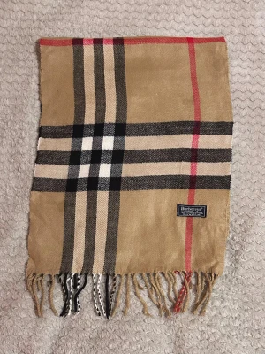 Beige rutig halsduk från Burberrys - Klassisk beige halsduk från Burberrys i 100% lammull med det ikoniska rutmönstret i svart, vitt och rött. Halsduken har fransar i båda ändar och en vävd etikett. Perfekt accessoar för att lyfta din outfit.