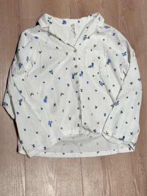 Vit pyjamaströja med blå blommor - Söt vit pyjamaströja med blå blommor och gröna blad. Skjortmodellen har knappar framtill, en bröstficka och lång ärm. 