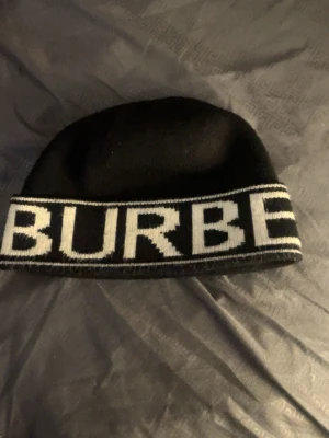 Svart mössa från Burberry - Säljer en svart mössa från Burberry med vit logotyptext runt hela kanten. Mössan är ribbstickad i nederkant och har en klassisk, enkel form. Perfekt för dig som vill ha en stilren accessoar med tydlig logga.