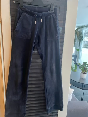 Mörkblå mjukisbyxor med snörning Juicy Couture  - Mörkblå mjukisbyxor i mjukt material med breda ben och snörning i midjan. Stora fickor fram och bakfickor med lock och knappdetaljer. Perfekta för chill dagar och riktigt sköna att ha på sig.
