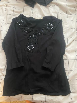 Svart topp med spets och pärlor - Svart topp med trekvartsärm, dekorerad med svart spets och glittriga pärlor framtill. Snygg detaljrik design som ger en unik look. Perfekt att styla med jeans eller kjol för en trendig outfit.