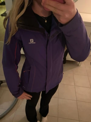 Skidjacka Salomon  - Säljer en lila skidjacka från Salomon med huva och röda detaljer längs ärmarna. Jackan har dragkedja framtill, justerbara ärmslut och två fickor med dragkedja samt en ficka för liftkort och fickor på insidan. Perfekt för kalla dagar och för skidsemester. Jackan är använd en skidsäsong och är i väldigt bra och fint skick. Kostar som ny på Salomon ca 3000 kr. Hör av dig om du har några frågor eller prisförslag❤️