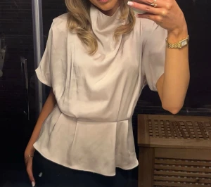 Ljusbeige satintopp - Snygg ljusbeige blus i satin med draperad krage och lös passform. Toppen har korta ärmar och markerad midja. Väl använd men i fint skick