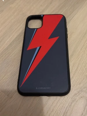 Svart mobilskal med röd blixt Bjornberry - Snyggt svart mobilskal från Bjornberry med en stor röd blixt och blå detalj på baksidan. Skalet har en stilren design och är gjort i ett tåligt syntetmaterial som skyddar mobilen mot repor och stötar. För iphone 11! Köpt för 250kr och är i princip ny skick. Säljer för att jag har skaffat en ny mobil💕