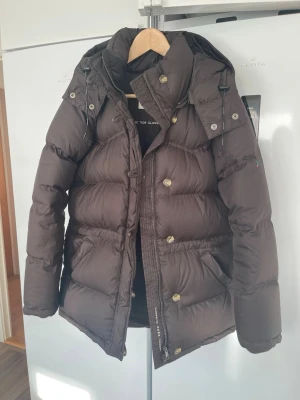 Brun dunjacka från Boomerang - Säljer en brun dunjacka från Boomerang, modell Authentic 7035 Classic Down Jacket. Jackan har avtagbar huva, dragkedja och knappar framtill samt flera fickor. Perfekt för kalla vinterdagar och riktigt skön att bära.