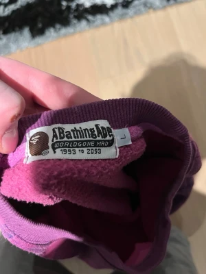 Lila hoodie från A Bathing Ape - Säljer en lila hoodie från A Bathing Ape med klassisk BAPE-logga på ärmen och insidan. Hoodien har ribbade muddar och är tillverkad i mjukt bomullsmaterial. Perfekt för dig som gillar streetwear och vill sticka ut med en ikonisk look.