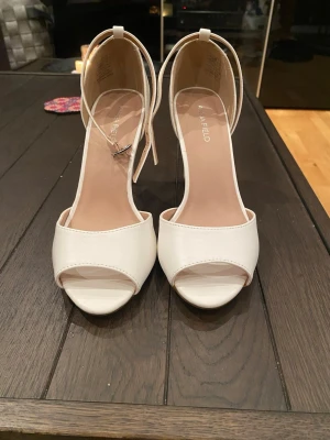 Vita höga klackar från Anna Field - Stilrena vita klackar från Anna Field med öppen tå och smal ankelrem med spänne. Skorna har hög klack och en elegant, minimalistisk design. Tillverkade i ett slätt material som ger en lyxig känsla. Perfekta för att lyfta vilken outfit som helst.