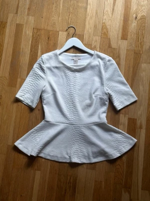 Vit peplumtop från H&M - Snygg vit peplumtop från H&M med strukturmönster i tyget. Toppen har rund halsringning och halvlånga ärmar. Den markerade midjan och utsvängda nederdelen ger en feminin siluett. Perfekt att styla med jeans eller kjol.