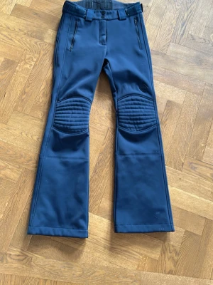 Mörkblå skidbyxor från J.Lindberg - Snygga mörkblå täckbyxor med quiltade förstärkningar över knäna och dragkedjeförsedda fickor framtill. Byxorna har utsvängda ben och bälteshällor i midjan. Perfekta för kalla dagar och outdoor-aktiviteter. Nypris ligger på 3200 och de är i nyskick förutom några små hål nere vid fötterna, se bild!