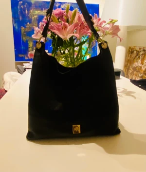 Svart axelremsväska från mulberry  - Stilren svart handväska i mjukt läder från mulberry. Väskan har en enkel axelrem med guldfärgade metalldetaljer och en liten guldfärgad logoplatta framtill. Perfekt för dig som gillar klassisk och tidlös design. Ny pris 12900kr