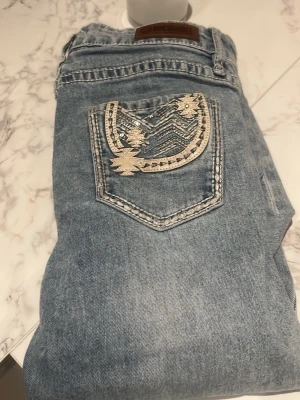 Ljusblå bootcut jeans med brodyr - Säljer ett par ljusblå jeans från rock and roll med bootcut passform. Jeansen har en snygg broderad bakficka med silverdetaljer och mönster. Klassisk femficksmodell och tillverkade i denim med en cool tvätt. Perfekta för dig som vill sticka ut lite extra. Storlek 16 men skulle säga w 23 elr w24 i jeans vanligtvis 