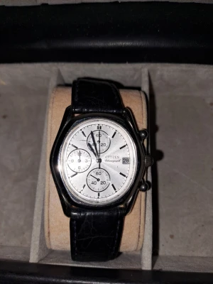 Citizen kronograf med svart läderband - Citizen kronografklocka med rund silverfärgad urtavla, tre subdials och datumvisning. Svart läderarmband med klassisk struktur och vit söm. Stilren design som passar till många looks.