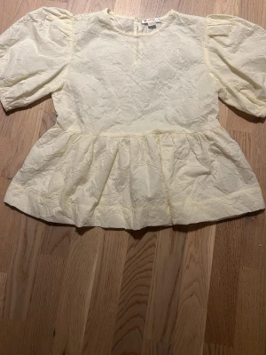 Gul topp med puffärm från Gina Tricot - Söt ljusgul topp från Gina Tricot med puffiga korta ärmar och strukturmönstrat tyg. Modellen har rund halsringning och volang nedtill som ger en peplum-effekt. Perfekt för dig som gillar en feminin och trendig stil.