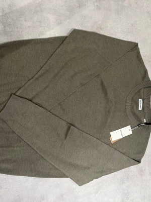 Olivgrön stickad tröja från Arket - Snygg olivgrön stickad tröja från Jack & Jones med rund halsringning och ribbade muddar vid ärmslut och nederkant. Tröjan har en clean och minimalistisk look, perfekt för lager på lager. Materialet känns mjukt och följsamt.