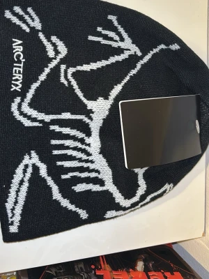 Svart mössa från Arc'teryx - Säljer en svart stickad mössa från Arc'teryx med vitt grafiskt mönster och logga på sidan. Mössan har en klassisk passform och är tillverkad i mjukt material som håller dig varm under kalla dagar. Perfekt för dig som gillar stilrena accessoarer.
