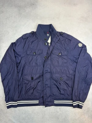 Blå vindjacka från Moncler - Snygg blå vindjacka från Moncler med fyra stora fickor framtill, ribbade muddar med vita ränder och hög krage. Jackan har både knappar och dragkedja samt en patch på ärmen. Perfekt för dig som vill ha en stilren och sportig look.