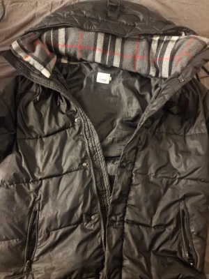 Svart pufferjacka Burberry England - Säljer en svart pufferjacka från Burberry England med huva och dragkedja. Jackan har två framfickor med dragkedja och ett rutigt foder i huvan med grå, svart, vit och röd färg. Märkeslogga på ärmen. Perfekt för kalla vinterdagar.