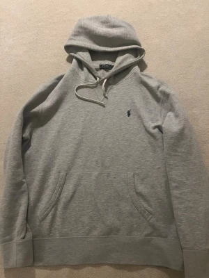 Grå hoodie från Polo Ralph Lauren - Snygg ljusgrå hoodie i L men sitter som M från Polo Ralph Lauren med klassisk huva och dragsko. Har en liten mörkblå logga broderad på bröstet och en stor magficka framtill. Tillverkad i mjukt bomullsmaterial som är perfekt för chill dagar.